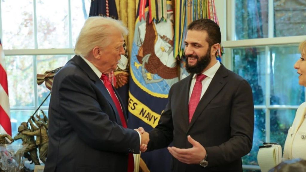 Trump and Al-Sharaa in dark suits shake hands inside an ornate office, with U.S. flags and a window behind them; a woman stands at the right edge of the frame.