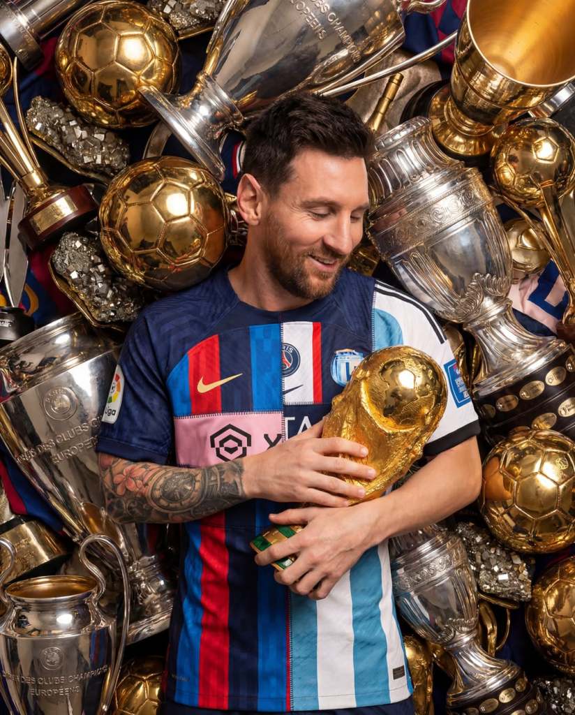 Lionel Messi holding the FIFA World Cup trophy in front of a display of various football trophies and awards.