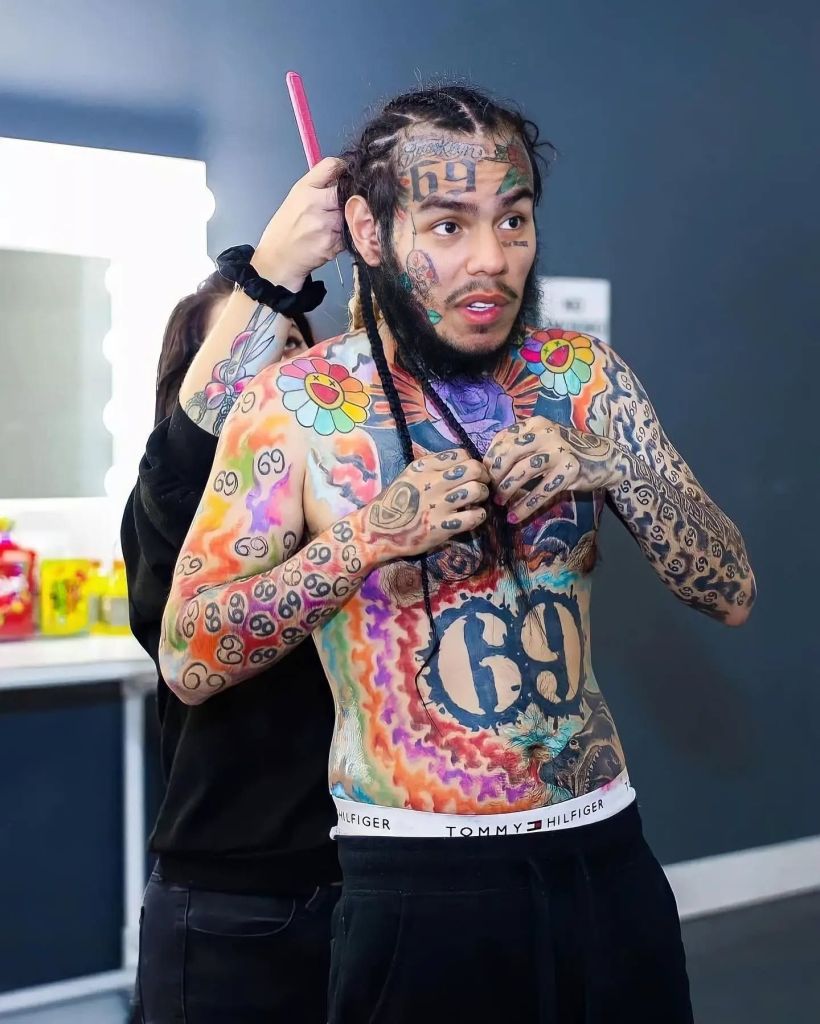 6ix9ine stands shirtless while someone braids his hair, showing his colourful full-torso tattoos, face tattoos, and patterned arm ink in a backstage room with bright lighting.