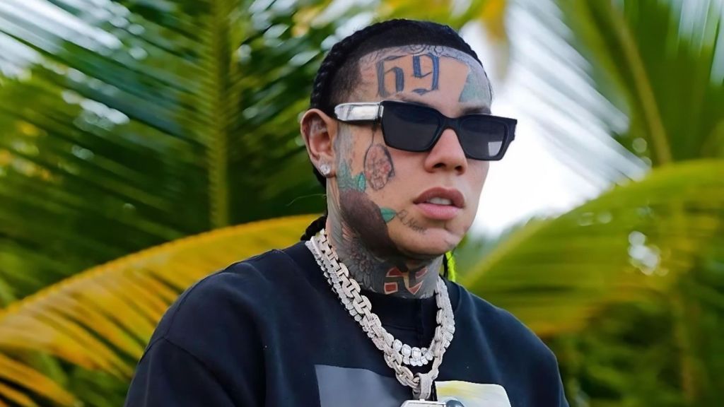Rapper 6ix9ine wearing dark sunglasses, braided hair, face tattoos, and layered diamond chains while standing outdoors in front of palm leaves.