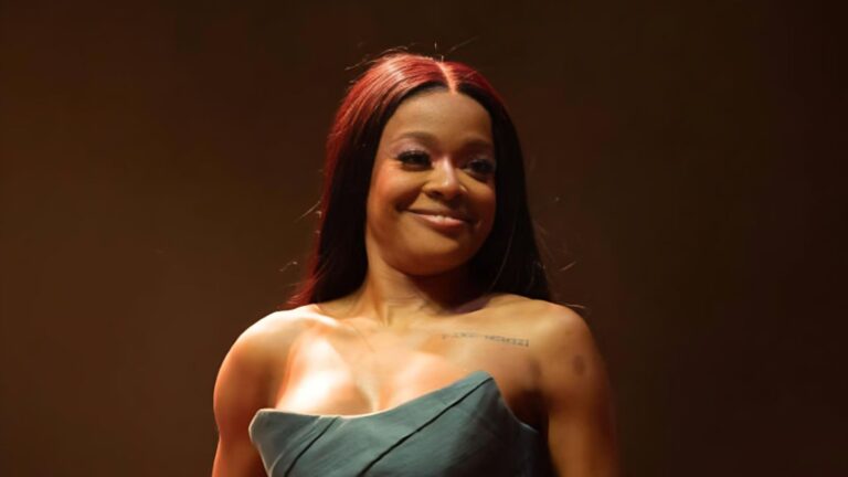 Azealia Banks smiling onstage while wearing a strapless green dress, with soft lighting highlighting her face and shoulders.