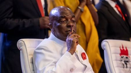 A close-up, seated portrait of Bishop David Oyedepo, the Presiding Bishop of the Living Faith Church Worldwide (Winners' Chapel). He is a dark-skinned, bald man wearing his signature white suit with a red lapel pin. He is looking down and to the side while holding his index finger to his lips in a gesture of silence, contemplation, or deep thought. He is seated in a white upholstered chair, and other attendees in dark and yellow clothing are visible in the background.