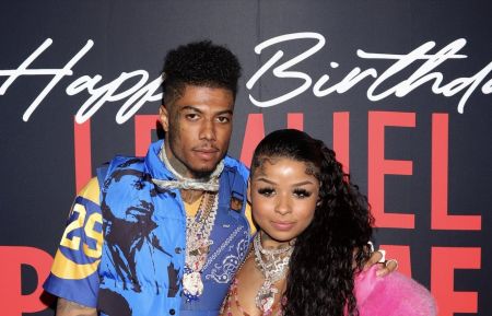 Photo of rappers Blueface (left: wearing a blue and yellow graphic jacket over a bandana print shirt, multiple diamond chains, and visible neck tattoos) and Chrisean Rock (right: long dark hair, heavy makeup, layered necklaces, and a pink fur sleeve) posing together in front of a black step-and-repeat backdrop with "Happy Birthday" and partial text "DANIEL PALACE".