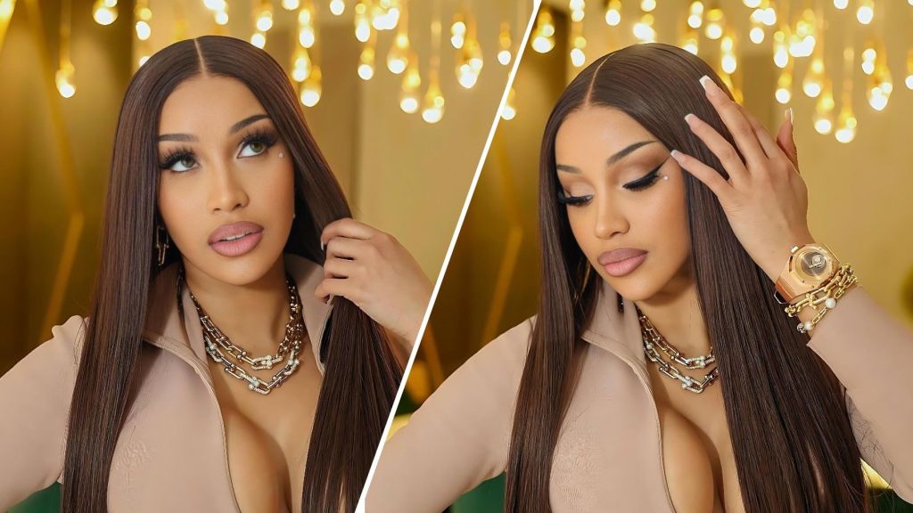 Cardi B poses indoors with long straight brown hair, wearing layered gold jewelry and a beige zip-front outfit as warm hanging lights glow in the background.
