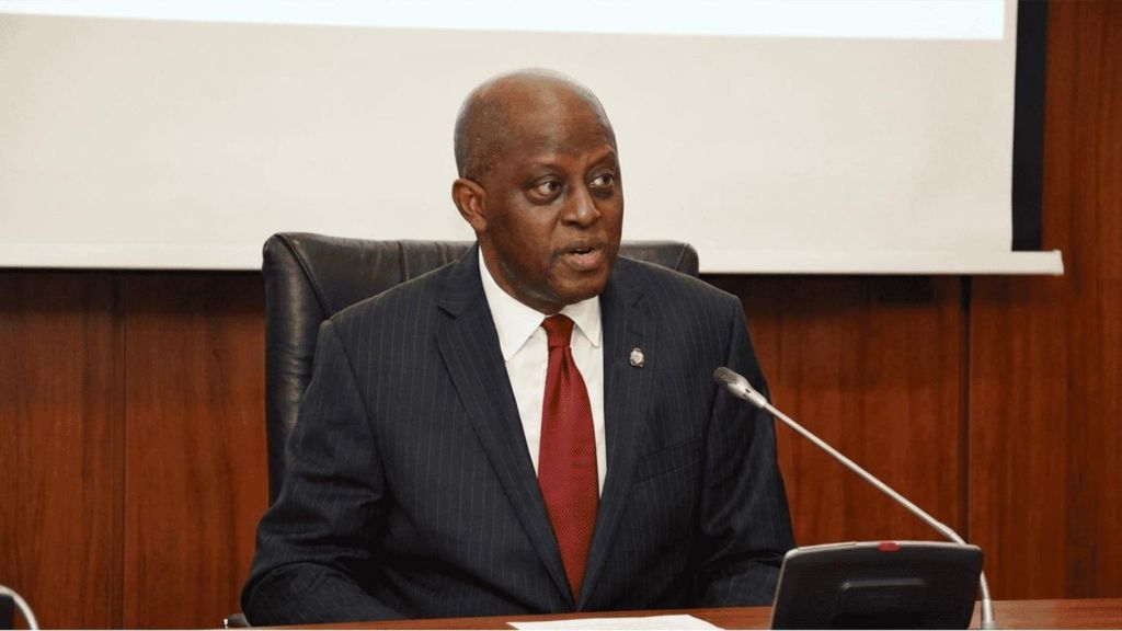 “Central Bank of Nigeria governor seated at a conference table, wearing a dark suit and red tie, speaking into a microphone during an official briefing