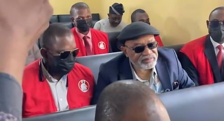 A photo showing former Minister Chris Ngige arriving at the Federal Capital Territory (FCT) High Court in Gwarinpa. Ngige is dressed in a blue suit and dark spectacles, walking alongside several EFCC officers in plain clothes and others wearing the commission's signature red tactical vests.