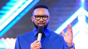 Nigerian pastor Dr. Chris Okafor preaching on stage, wearing a blue suit and glasses, holding a microphone in one hand while raising the other palm open, against a vibrant blue and purple lit backdrop with abstract light patterns.