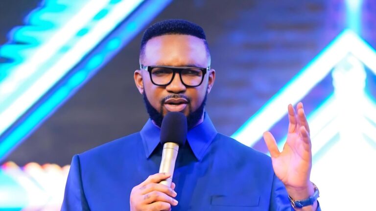 Nigerian pastor Dr. Chris Okafor preaching on stage, wearing a blue suit and glasses, holding a microphone in one hand while raising the other palm open, against a vibrant blue and purple lit backdrop with abstract light patterns.