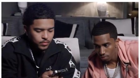 Photo of Sean "Diddy" Combs' sons Christian "King" Combs (left: wearing a black jacket with white patterned sleeves and "SAINT" text, holding a phone) and Justin Combs (right: in a pink jacket over a white shirt with multiple silver chains) sitting side-by-side on a couch in a dimly lit room, both looking down with serious expressions.