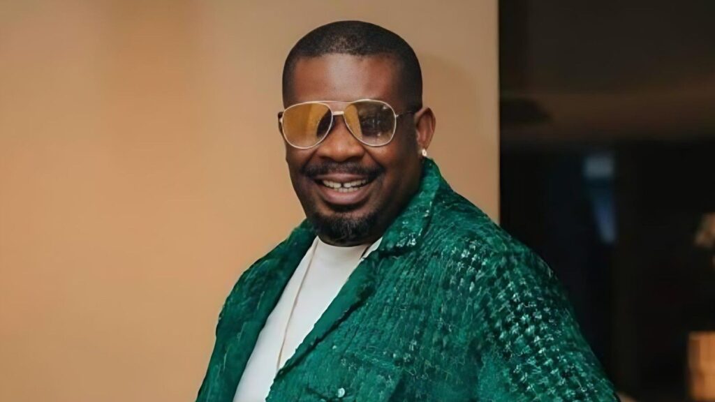 Portrait of Nigerian music producer and entrepreneur Don Jazzy, smiling broadly while wearing tinted aviator sunglasses, a white shirt, and a textured emerald green jacket, with a pearl earring visible, against a neutral indoor background.