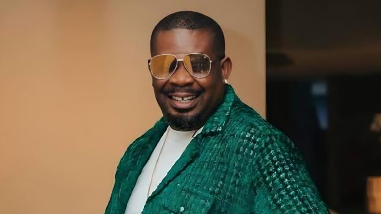 Portrait of Nigerian music producer and entrepreneur Don Jazzy, smiling broadly while wearing tinted aviator sunglasses, a white shirt, and a textured emerald green jacket, with a pearl earring visible, against a neutral indoor background.