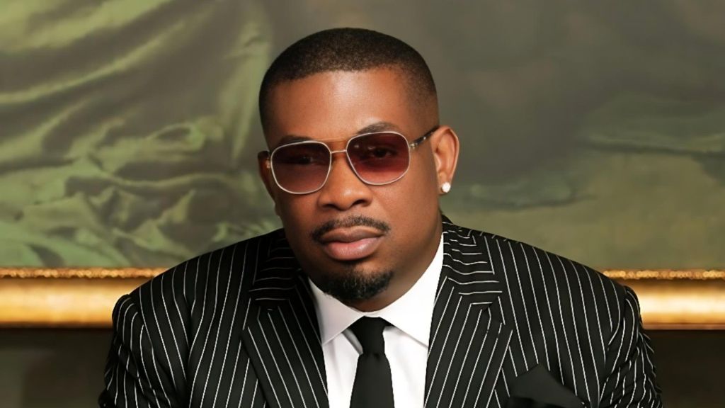 Portrait of Nigerian music producer and Mavin Records CEO Don Jazzy, wearing a black pinstripe suit, white dress shirt, black tie, tinted aviator-style sunglasses, and a pearl earring, seated against a classical painting backdrop.