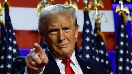 Portrait of U.S. President Donald Trump at a formal event, wearing a dark navy suit, white dress shirt, red tie, and American flag lapel pin, pointing directly at the camera with a serious expression against a backdrop of American flags and golden eagle emblems.
