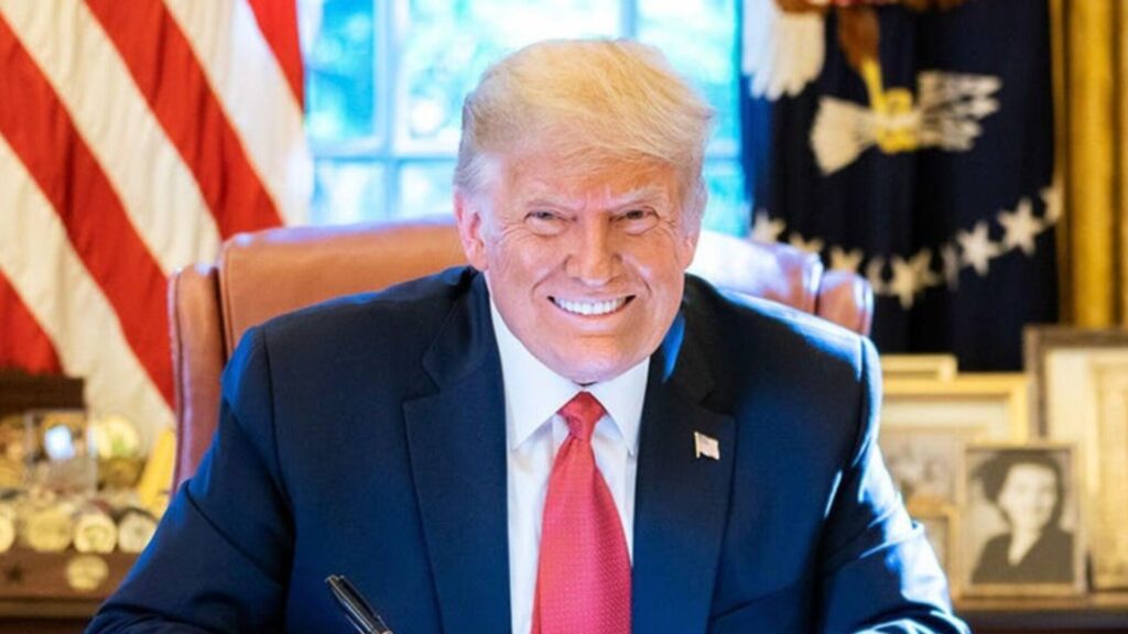 A smiling Donald Trump sits at a desk in an office setting, wearing a dark suit and red tie, with an American flag and framed photos visible in the background.