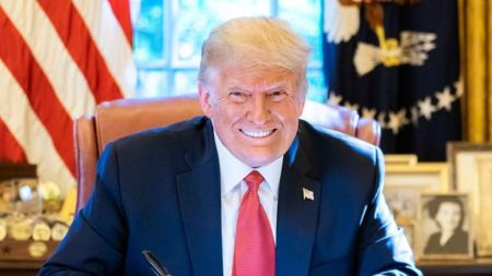 A smiling Donald Trump sits at a desk in an office setting, wearing a dark suit and red tie, with an American flag and framed photos visible in the background.