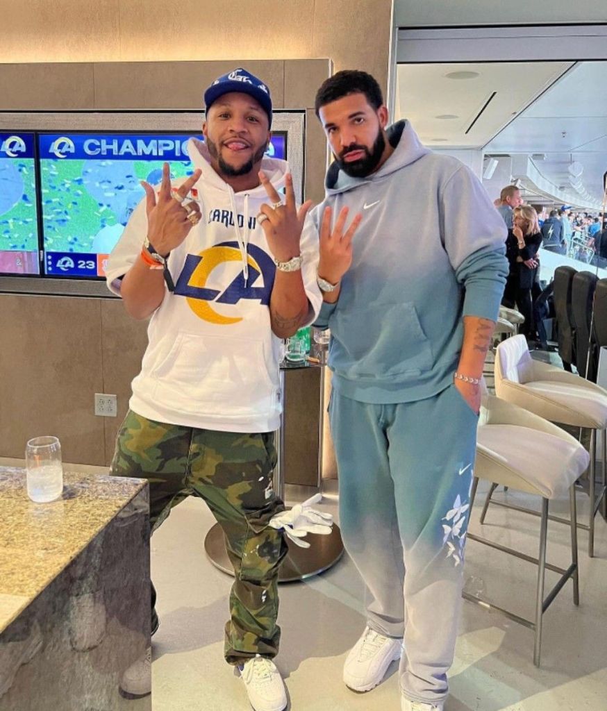 Photo of American streamer and content creator BenDaDonnn (left, wearing a white Los Angeles Rams hoodie, blue cap, camouflage cargo pants, and multiple rings while flashing peace signs) posing with Canadian rapper Drake (right, in a gradient gray Nike hoodie and matching sweatpants, also flashing a peace sign) in a luxury suite, with a TV screen showing an LA Rams championship celebration in the background.