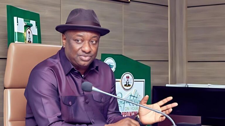 Festus Keyamo sits at a government briefing desk, speaking into a microphone while gesturing with one hand.