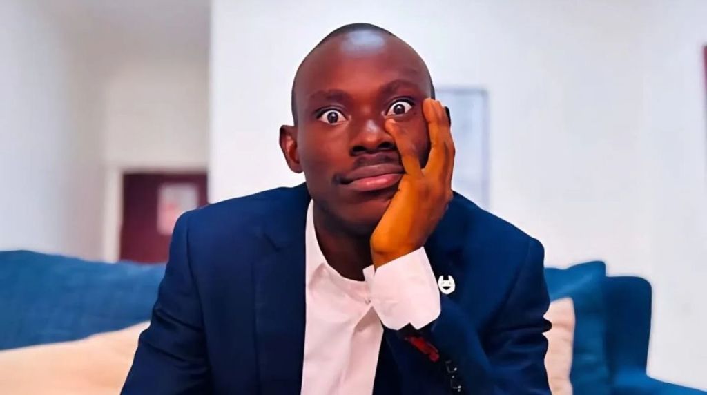 “A close-up photo of GehGeh wearing a navy blue suit, resting his face on one hand, with wide eyes and a neutral expression while seated indoors.
