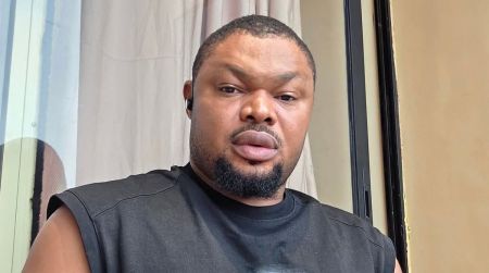 Harrison Gwamnishu looking into the camera while sitting indoors, wearing a sleeveless black shirt with a curtain and wall behind him.