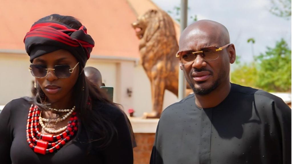 2Baba stands beside Natasha Osawaru outdoors, both wearing dark outfits and sunglasses, with a large lion statue visible in the background.