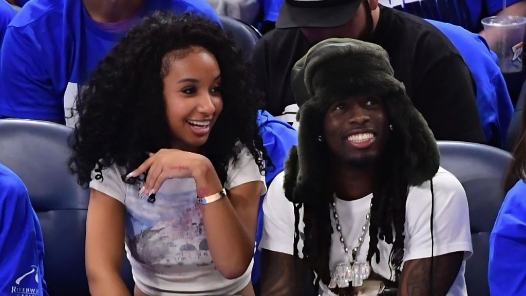 Photo of American streamer Kai Cenat (right: long dreadlocks with a large black furry hat, white T-shirt, silver chains, and visible tattoos, smiling) sitting courtside at a basketball game next to Gabrielle Alayah (left: curly dark hair, white graphic T-shirt, bracelet, smiling with hand near chin), surrounded by spectators in blue shirts.