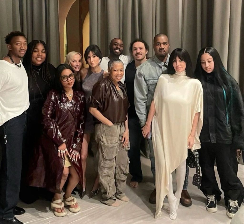 Group photo of Kanye West (Ye) and his extended family and friends indoors against gray curtains—front row (left to right): North West in a shiny maroon coat and platform sandals, Bianca Censori in loose beige cargo pants and brown leather top barefoot, Kim Kardashian in an asymmetrical cream silk dress; back row includes Saint West, Chicago West, Khloé Kardashian, Kris Jenner, Psalm West, Kanye West in denim shirt, and others smiling together.