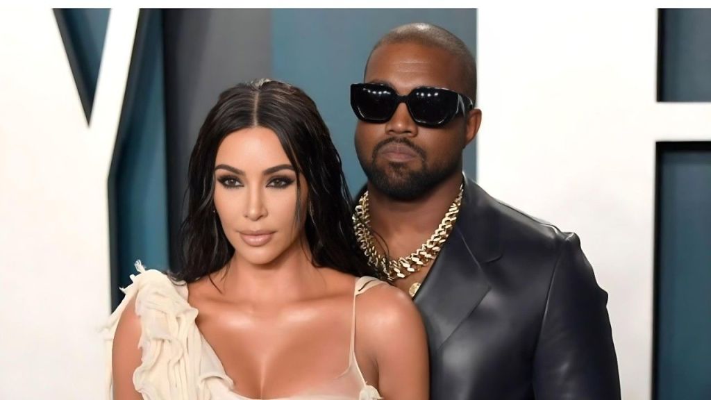 Photo of Kim Kardashian (left: wearing a cream-colored off-shoulder gown with ruffled detailing and loose wavy dark hair) and Kanye West (right: in a black leather jacket, large dark sunglasses, and multiple thick gold chains) posing together at a formal event against a white backdrop with partial "V" lettering (likely Vanity Fair Oscar Party).