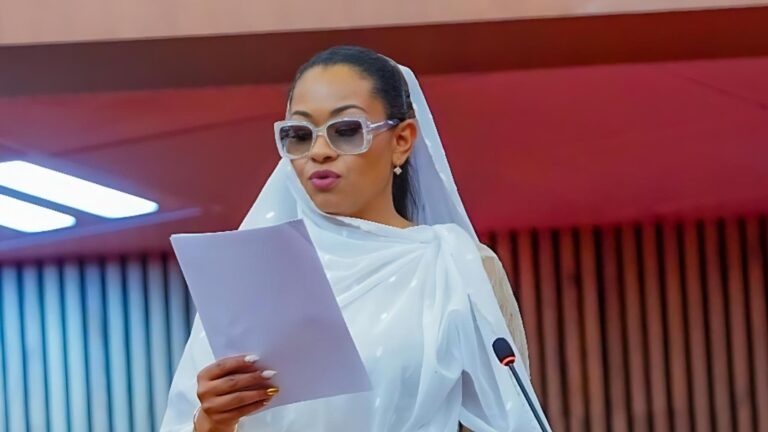 Natasha Akpoti dressed in a white outfit and translucent veil stands at a podium, reading from a sheet of paper while wearing large clear-framed glasses.
