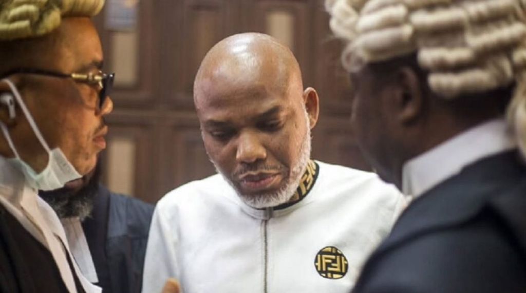 Nnamdi Kanu stands in court wearing a white outfit as he speaks with two of his lawyers, who are dressed in legal wigs and gowns.