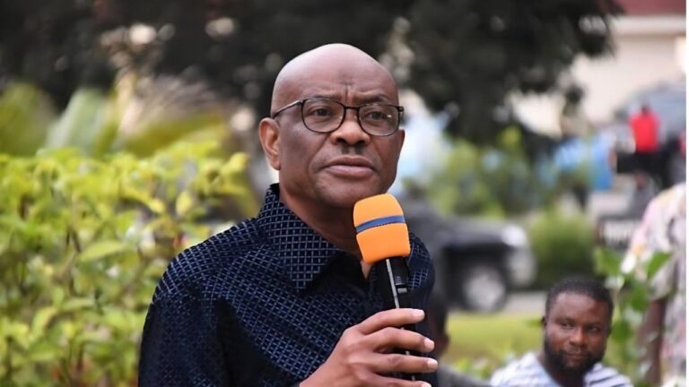 Nigerian politician and current Minister of the Federal Capital Territory, Nyesom Wike, speaking outdoors into a yellow-and-blue microphone while wearing glasses and a dark patterned shirt, with blurred greenery and people in the background.