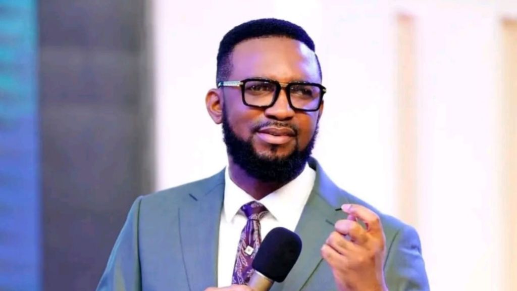 Portrait of Nigerian pastor and founder of Mountain of Liberation and Miracle Ministry, Dr. Chris Okafor, speaking into a microphone while wearing a light gray suit, white dress shirt, patterned purple tie, and large clear-framed glasses, against a blurred indoor stage background with colorful lighting.