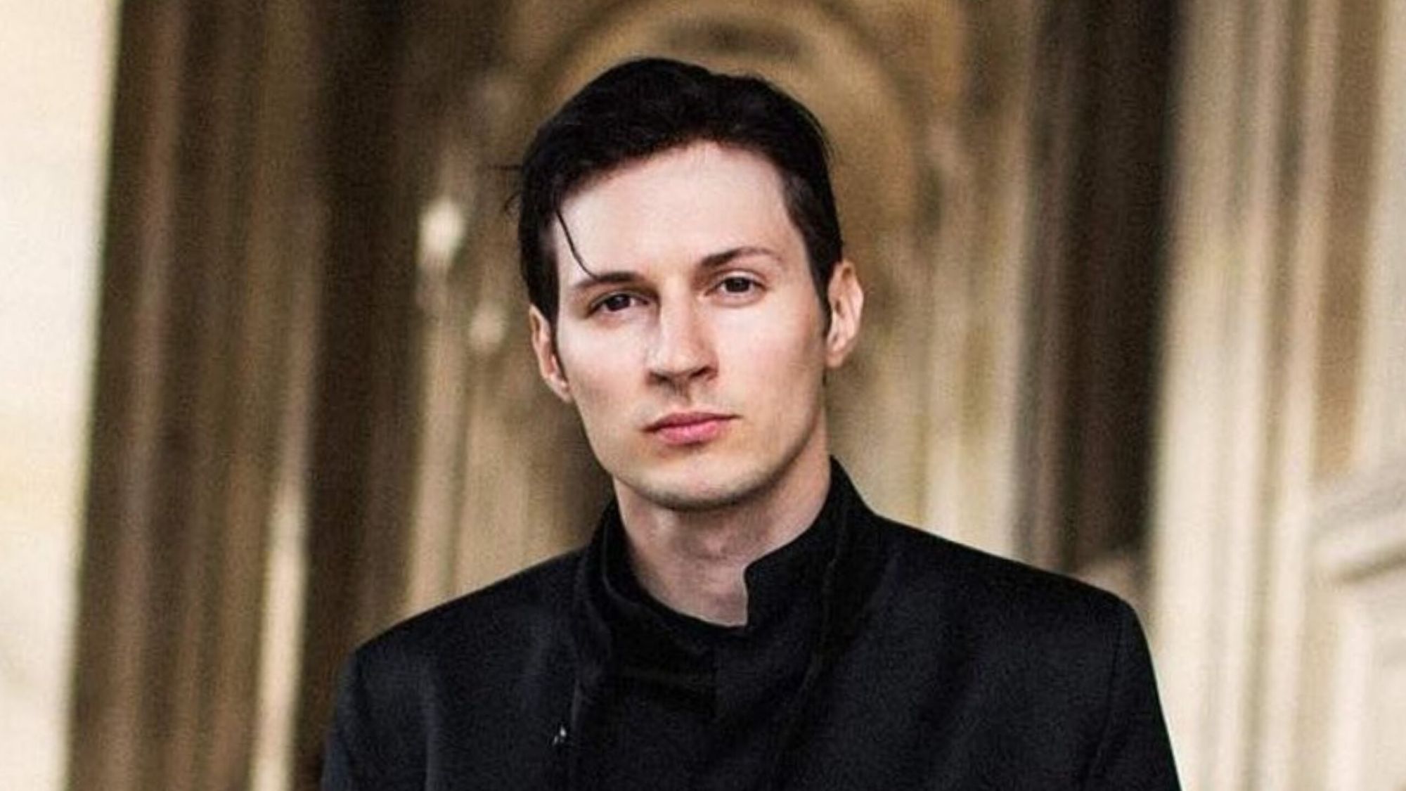 Portrait of Pavel Durov, founder and CEO of Telegram, wearing a black high-collar jacket, looking directly at the camera with a serious expression against a blurred classical architectural background with columns.