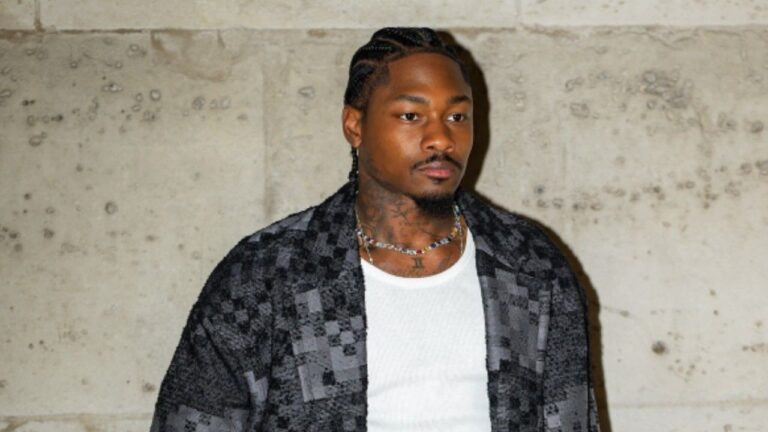 Portrait of American NFL wide receiver Stefon Diggs, wearing a white tank top under an open black-and-gray patterned jacket, with braided cornrows, visible neck and chest tattoos, and a multicolored beaded necklace, standing against a textured concrete wall background.
