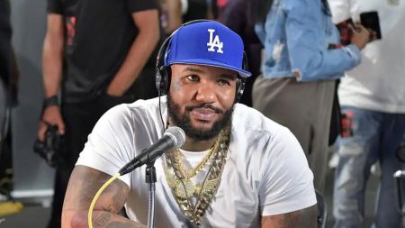 A close-up, chest-up shot of American rapper The Game (Jayceon Terrell Taylor) sitting in front of a microphone. He is a muscular Black man with a full beard, wearing a white t-shirt, a blue baseball cap with a white "LA" logo (Los Angeles Dodgers), over-the-ear headphones, and multiple thick gold chains around his neck. He appears to be recording or participating in a podcast or radio show.