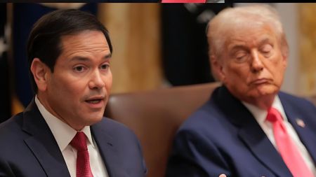 A public official speaks during a formal meeting while the U.S. president, seated beside him in a dark suit and pink tie, appears to have his eyes closed and head tilted downward as if resting.