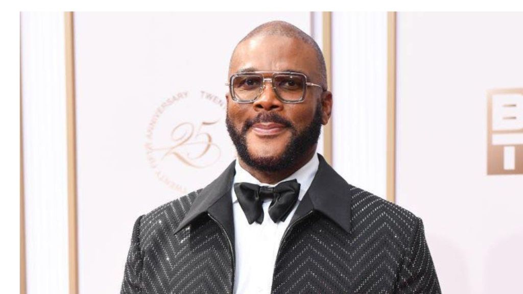 American actor, filmmaker, and producer Tyler Perry at a formal event, wearing a black sequined chevron-patterned jacket over a white dress shirt and black bow tie, with large clear-framed glasses, against a white backdrop featuring "25th Anniversary" text.