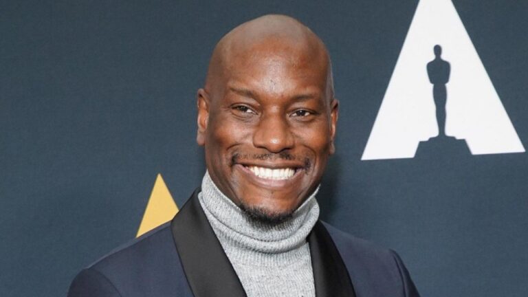 American actor and singer Tyrese Gibson smiling at a formal event, wearing a dark suit jacket with black lapels over a light gray turtleneck sweater, against a dark backdrop featuring the white Oscar statuette silhouette and a golden triangle element.