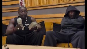 Photo of Nigerian social media activist VeryDarkMan (left: muscular build, braided hair, wearing a black sleeveless outfit and beaded necklaces, holding stacks of cash while seated) and Grammy-winning singer Burna Boy (right: in a black hooded tracksuit with arms crossed, looking serious) sitting side-by-side on a yellow couch in a studio or lounge setting with wooden paneling.