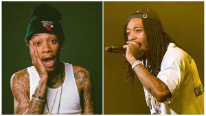 A portrait of rapper Wiz Khalifa, seen performing on stage during a concert in late 2025. He is shirtless, displaying his extensive collection of tattoos across his torso and arms, and is wearing multiple layered diamond chains. He has his signature dreadlocks tied back and is holding a microphone while smiling at the audience under bright stage lights.