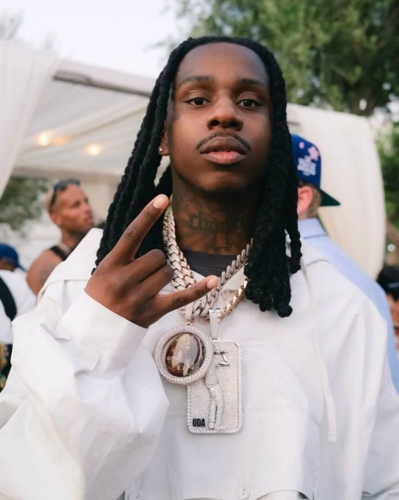 Polo G poses outdoors wearing a white jacket, layered diamond chains and a large iced-out pendant, holding up a hand gesture while looking directly at the camera. Other people and a tented setup appear blurred in the background.