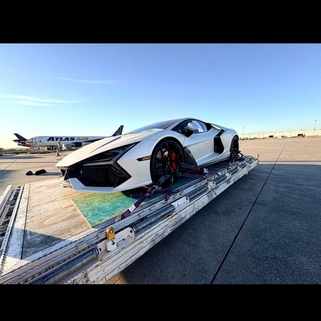 A white 2026 Lamborghini Revuelto secured on a cargo pallet at an airport, with an airplane in the background.