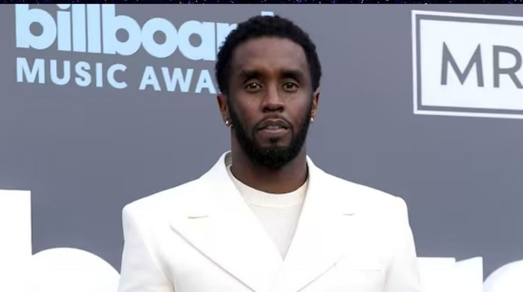 Sean "Diddy" Combs posing on the red carpet at the 2022 Billboard Music Awards in Las Vegas. He is wearing a crisp white suit with a matching white t-shirt and small hoop earrings, standing against a grey backdrop featuring the Billboard Music Awards logo.