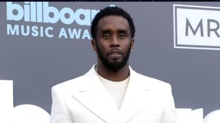 Sean "Diddy" Combs posing on the red carpet at the 2022 Billboard Music Awards in Las Vegas. He is wearing a crisp white suit with a matching white t-shirt and small hoop earrings, standing against a grey backdrop featuring the Billboard Music Awards logo.