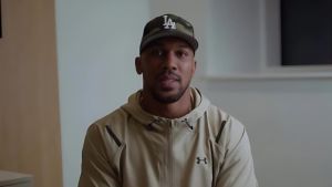 Anthony Joshua sits facing the camera wearing a beige Under Armour hoodie and camouflage LA baseball cap. He has a somber expression as he delivers a video message about the loss of his friends, Latz and Sina.