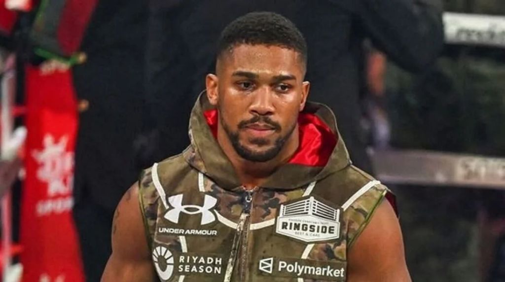 Anthony Joshua stands in a boxing ring, looking downwards with a serious expression. He is wearing a camouflage-patterned sleeveless hoodie featuring sponsorship logos for "Under Armour," "Riyadh Season," and "Polymarket." The background is blurred, showing ring ropes and stadium lighting.