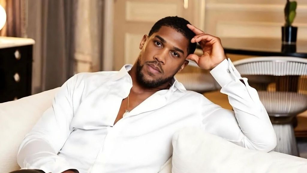 British heavyweight boxer Anthony Joshua sitting on a white sofa, wearing a crisp white unbuttoned shirt and black trousers. He is leaning back comfortably with his left hand resting against his temple, looking directly at the camera with a calm, serious expression. A blurred luxury interior with a lamp and curtains is visible in the background.