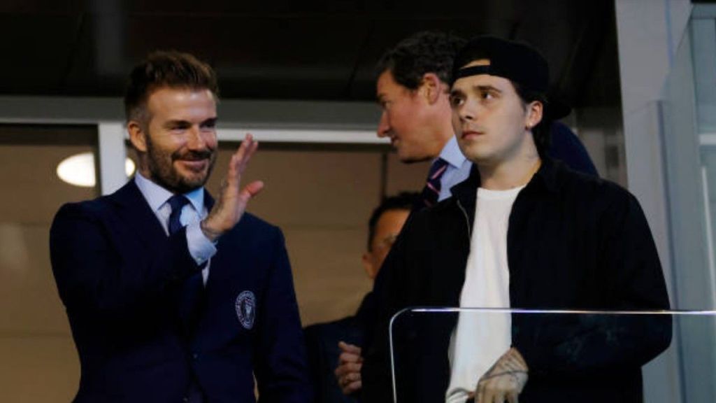 David Beckham, wearing a navy suit with an Inter Miami crest, waves to the crowd from a stadium box while standing next to his son Brooklyn Beckham, who is wearing a black jacket, white t-shirt, and a backwards black baseball cap.