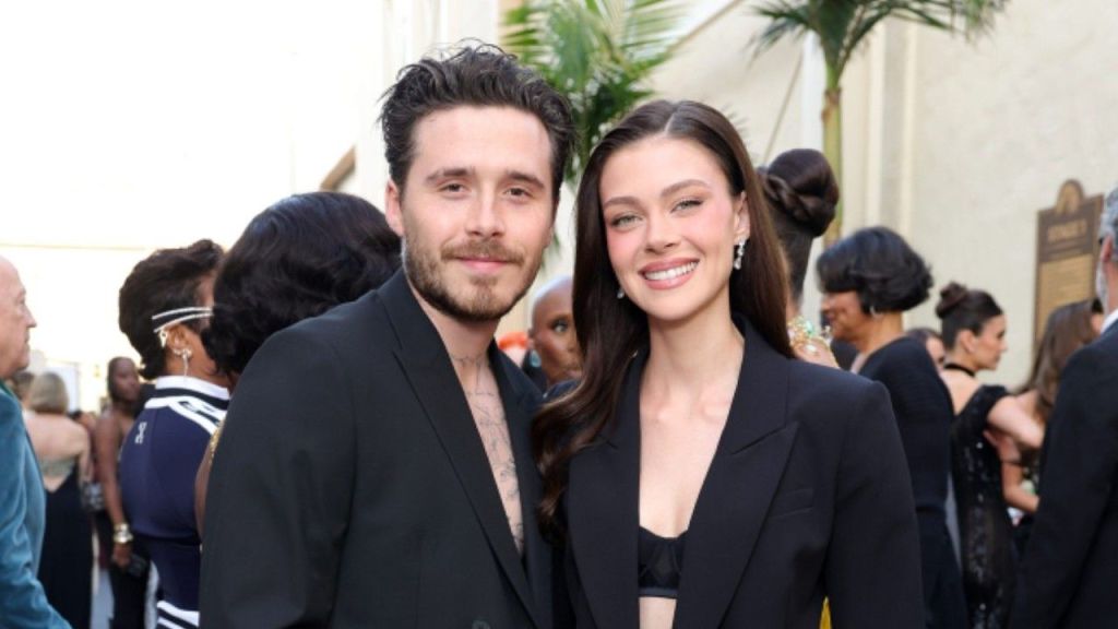 A medium shot of Brooklyn Beckham and Nicola Peltz posing together at the 3rd Annual Academy Museum Gala in 2023. Brooklyn (left) wears a black suit jacket with no shirt underneath, revealing his chest tattoos, while Nicola (right) wears a matching black blazer and skirt. They are both smiling warmly at the camera.