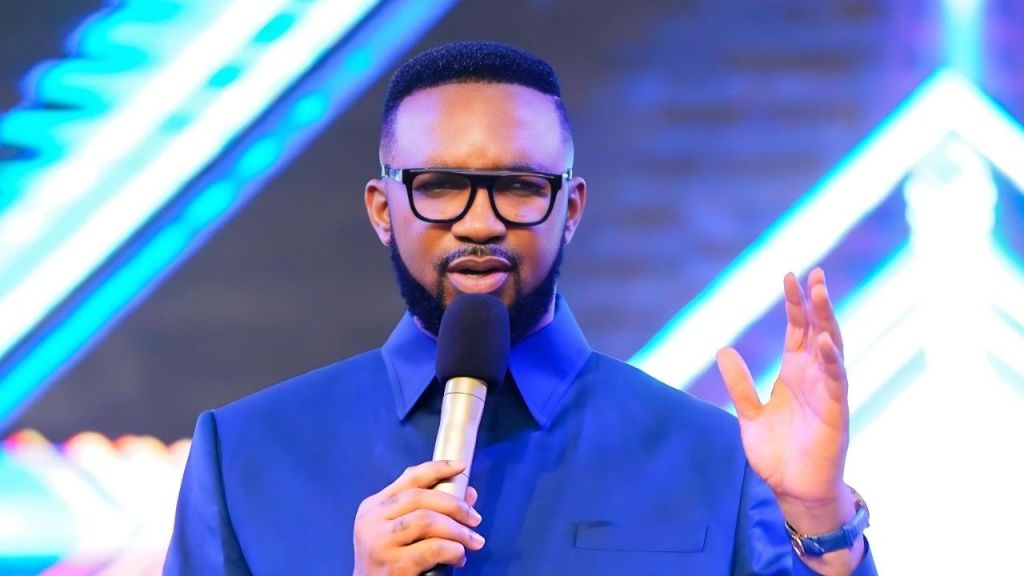 Pastor Chris Okafor of Mountain of Liberation and Miracles Ministries speaking into a microphone while wearing a blue suit and glasses against a neon blue background.