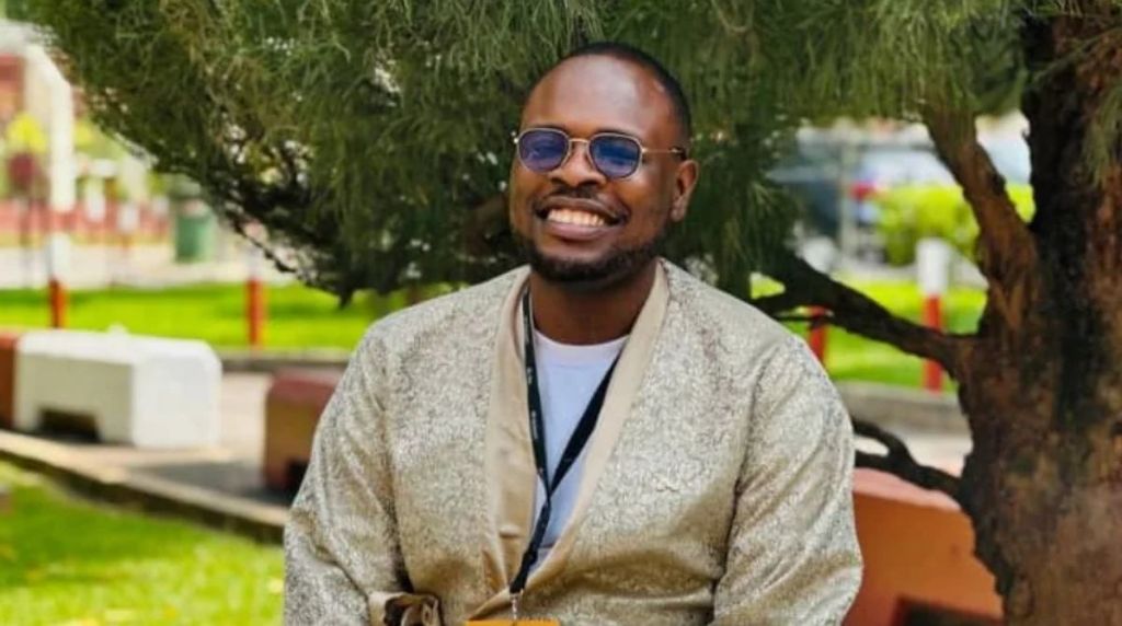 Media personality Chude Jideonwo smiling broadly while sitting outdoors, wearing blue-tinted sunglasses and a textured beige outfit with a lanyard around his neck.